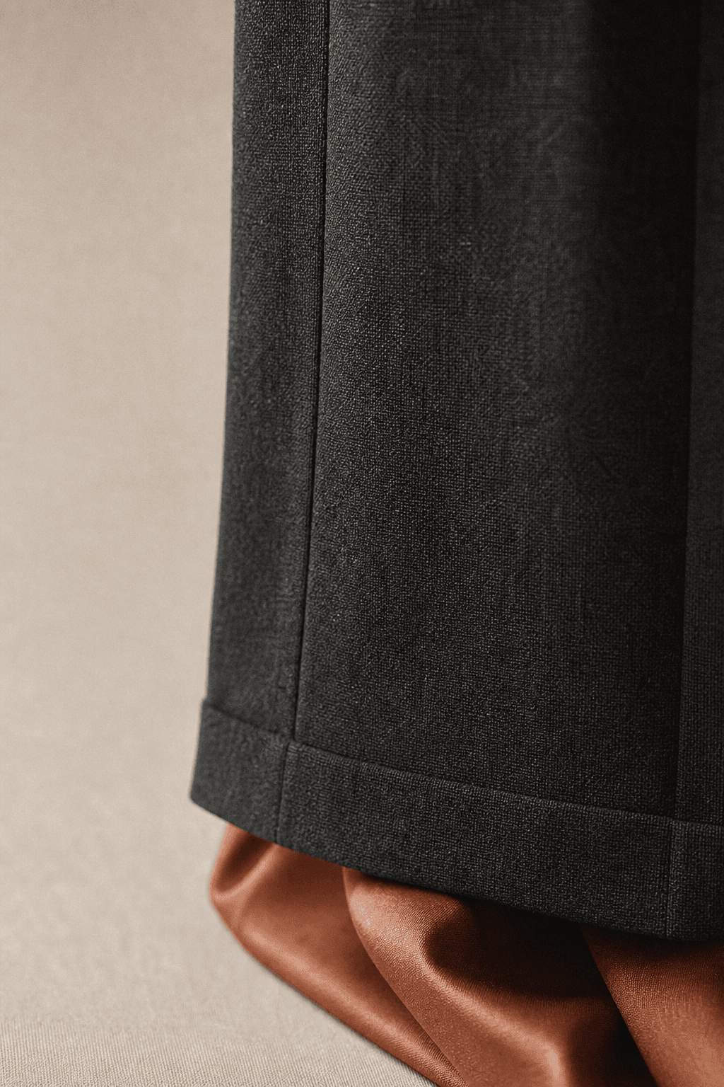 Close-up of the pressed hem and side seam of charcoal wool trousers resting over a fold of terracotta silk.